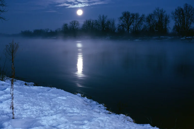 Full moon reflecting on still winter river at night with snow-covered bank and mist rising from water