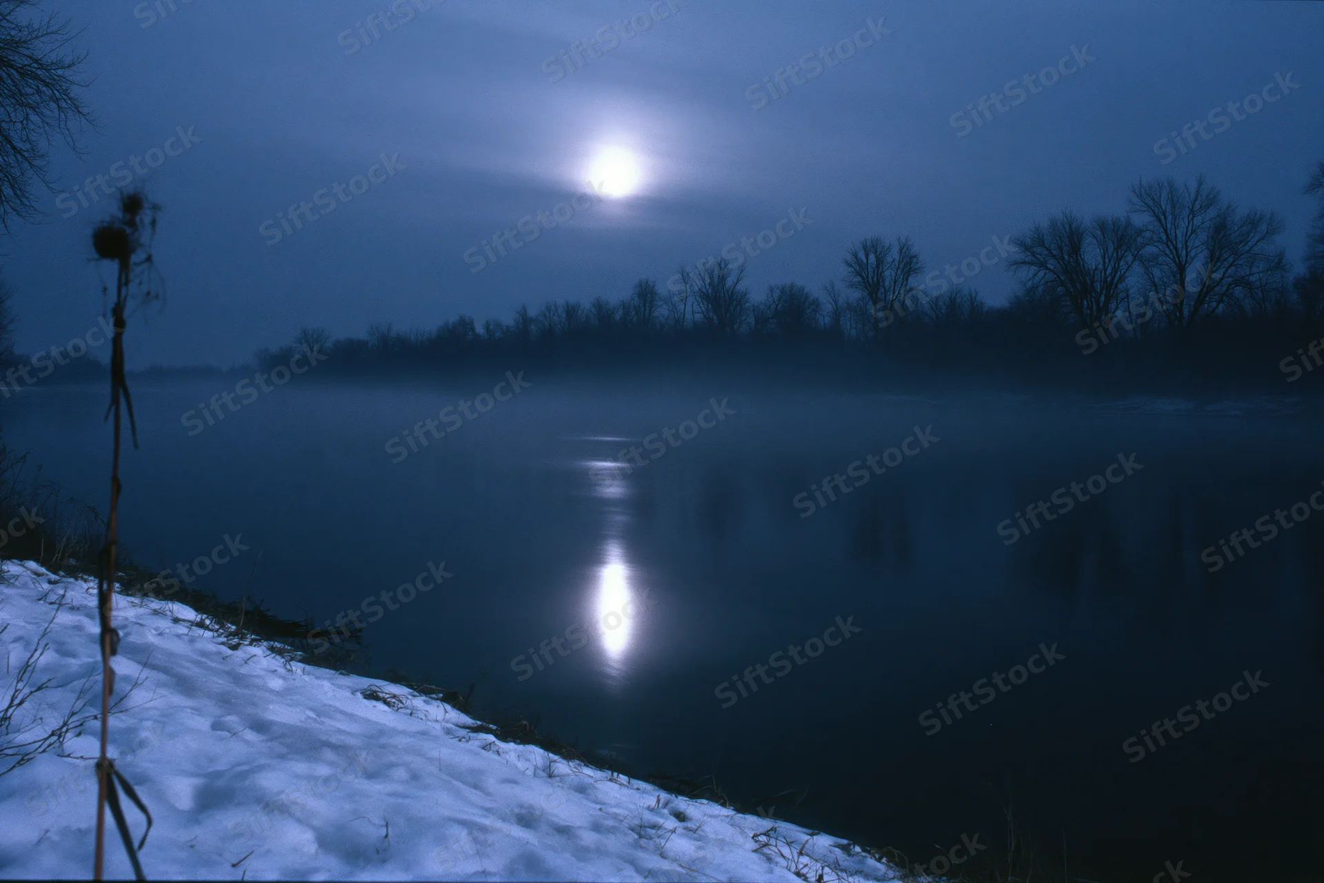 Full moon reflecting on a still winter river at night with mist rising from the water and snow on the bank