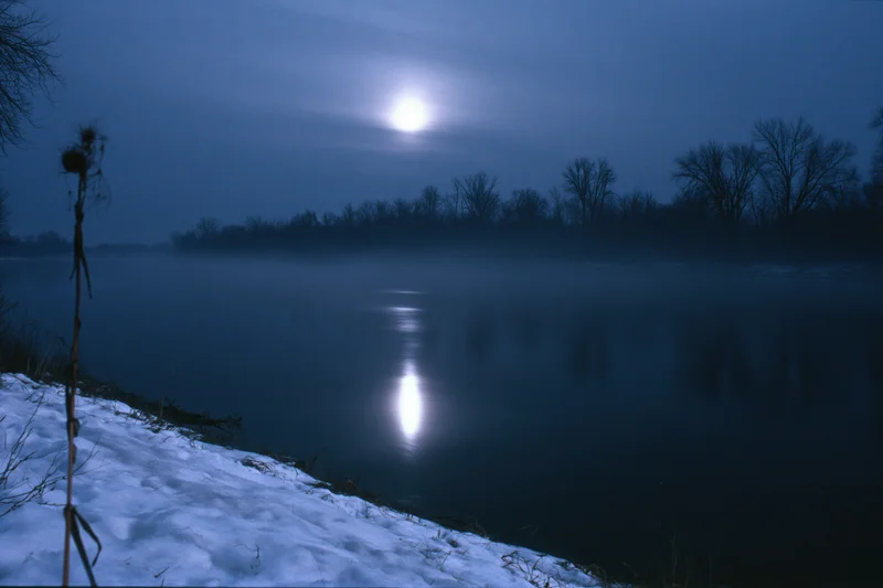 Full moon reflecting on a still winter river at night with mist rising from the water and snow on the bank