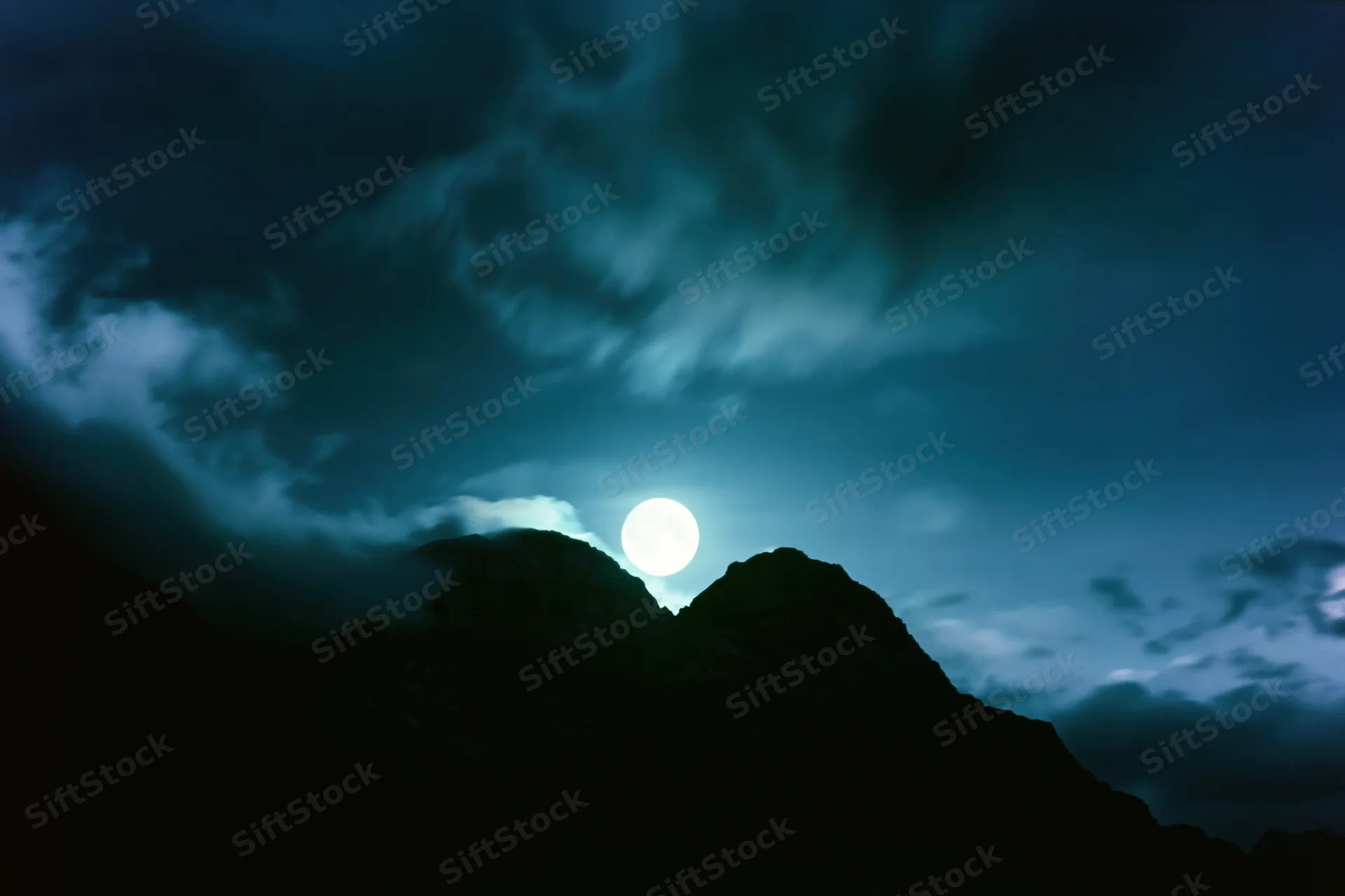 Full moon rising over dark mountain silhouettes in a cloudy night sky