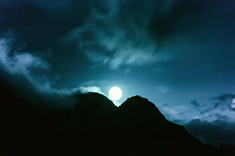 Full moon rising over dark mountain silhouettes in a cloudy night sky