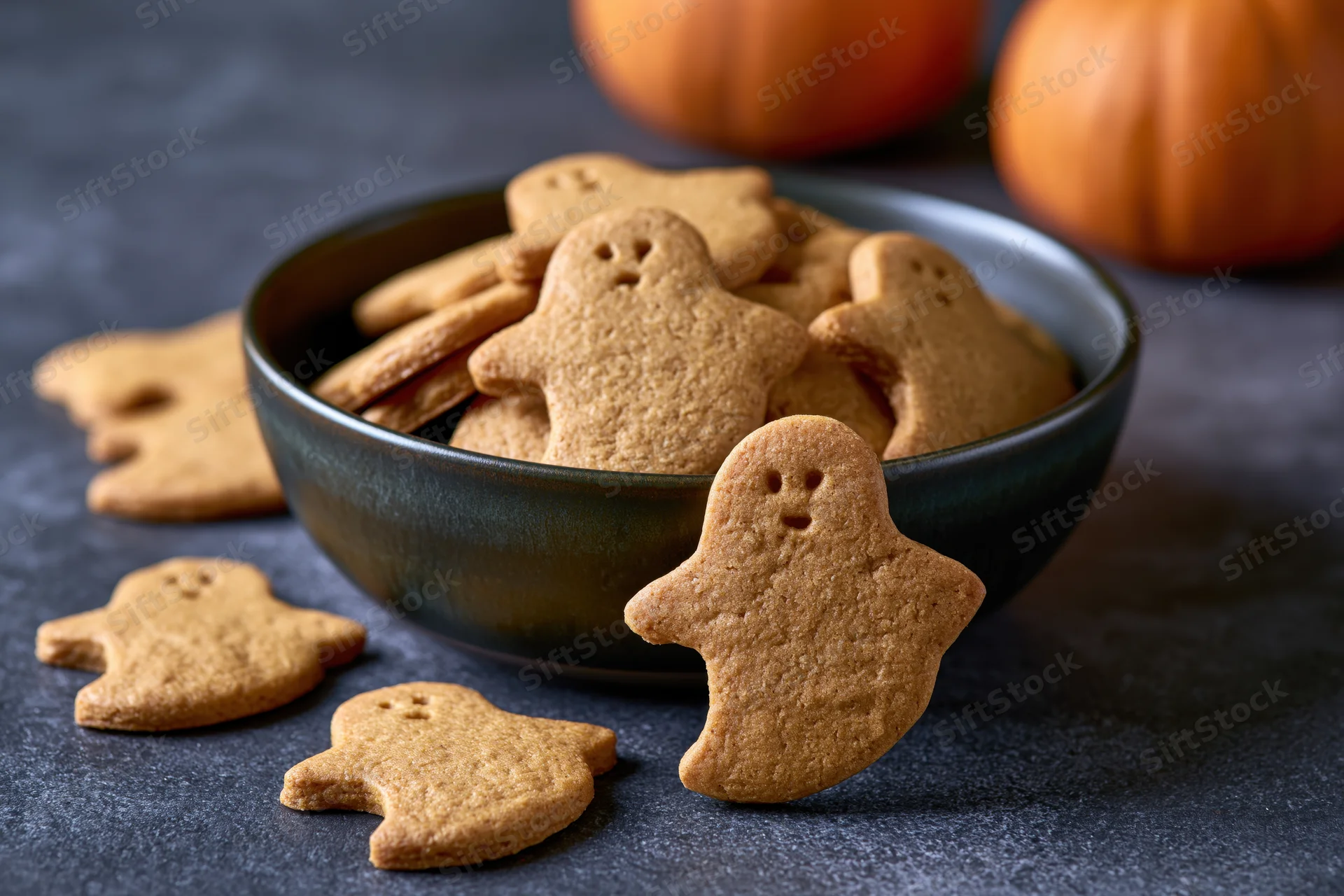 Ghost-shaped pumpkin cookies in a dark ceramic bowl on a dark surface with pumpkins in the background