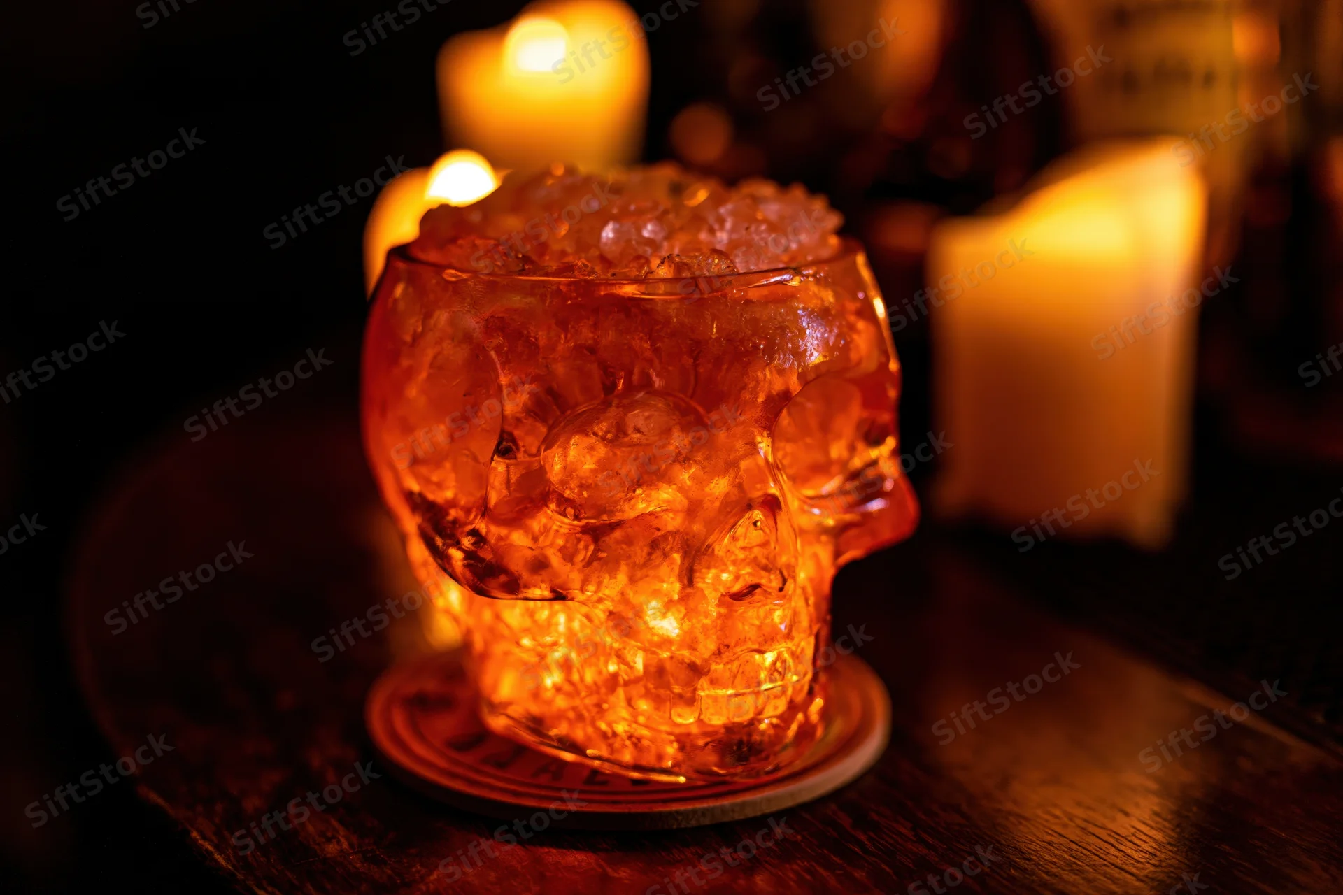 Glass skull cocktail cup filled with crushed ice and amber drink on wooden table with candles glowing in background