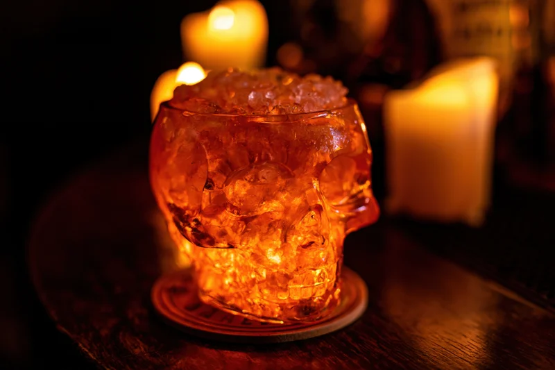 Glass skull cocktail cup filled with crushed ice and amber drink on wooden table with candles glowing in background