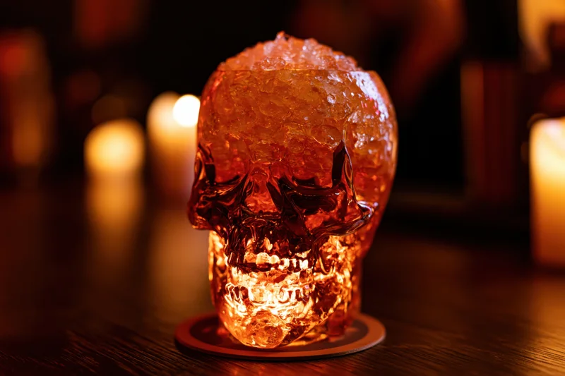 Glass skull cocktail cup filled with crushed ice and amber drink on wooden bar table with candles glowing in background