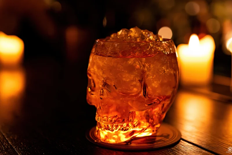 Skull-shaped glass filled with crushed ice and amber drink on wooden table with candles glowing in background