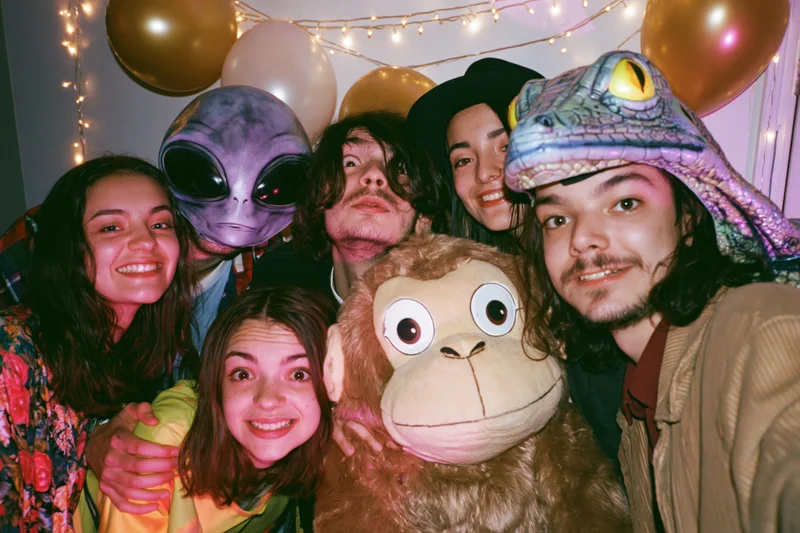 Five young adults posing together at an indoor costume party with masks and balloons