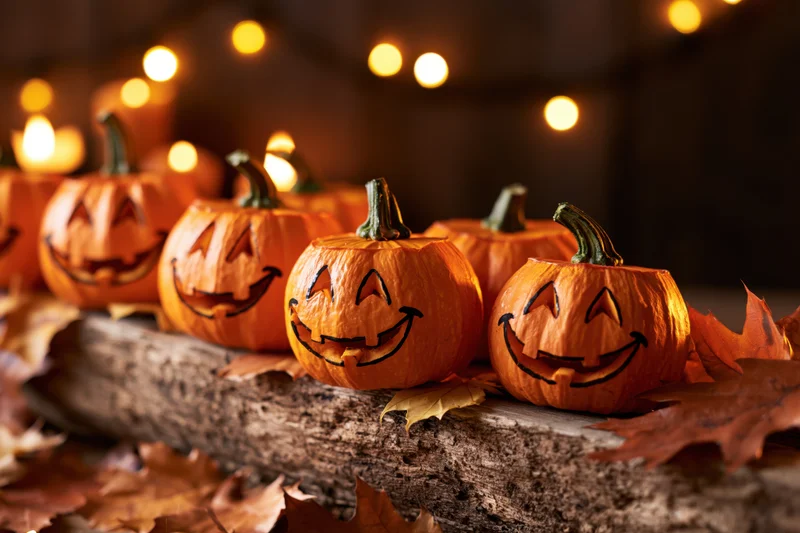 Carved pumpkins with smiling faces arranged on a wooden log surrounded by autumn leaves and warm string lights