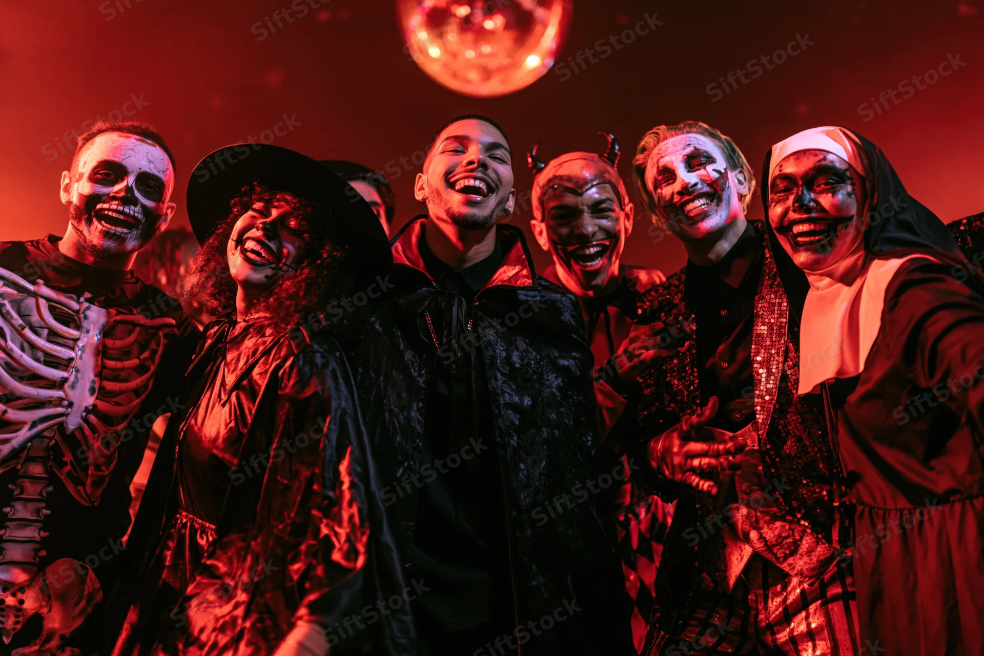 Group of young adults in halloween costumes laughing and dancing at a party under red disco lighting