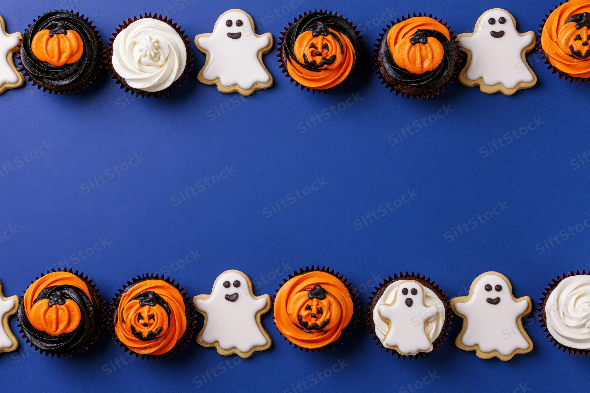 Halloween cupcakes and ghost cookies arranged in two rows on blue background with copyspace in center