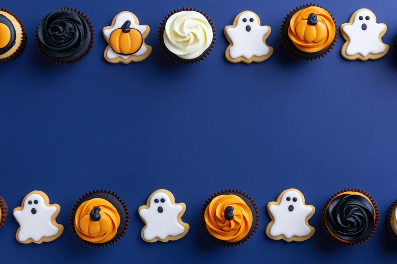 Halloween-themed cupcakes and ghost-shaped cookies arranged in two rows on a blue background with copyspace in the center