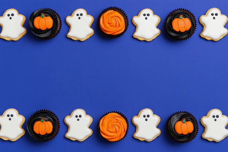 Ghost-shaped cookies and pumpkin-decorated cupcakes arranged in two rows on a blue background