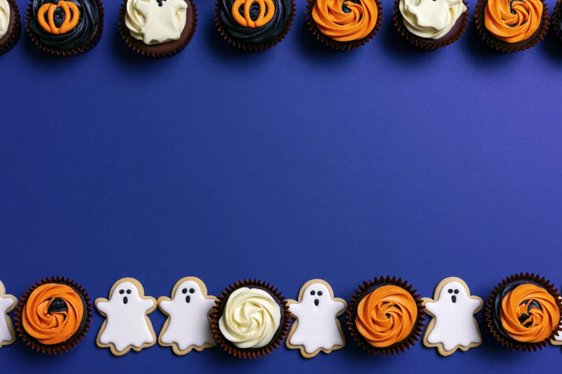 Halloween-themed cupcakes and ghost cookies arranged in rows on blue background with copyspace
