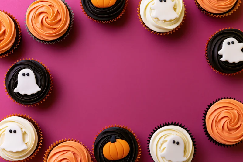 Halloween cupcakes with ghost and pumpkin decorations arranged in a border on a purple surface