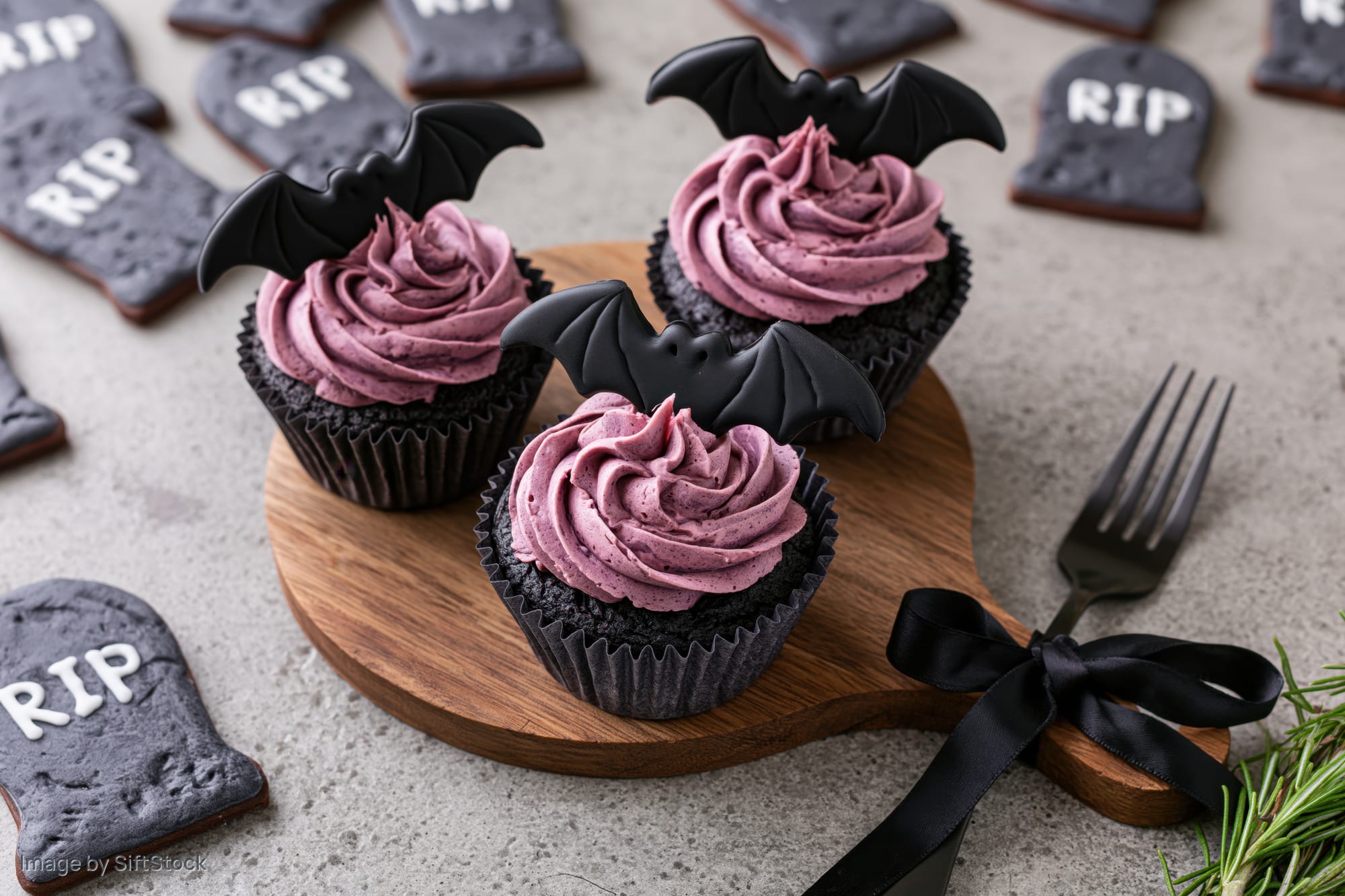Three halloween cupcakes with purple frosting and bat toppers on a wooden board surrounded by tombstone cookies
