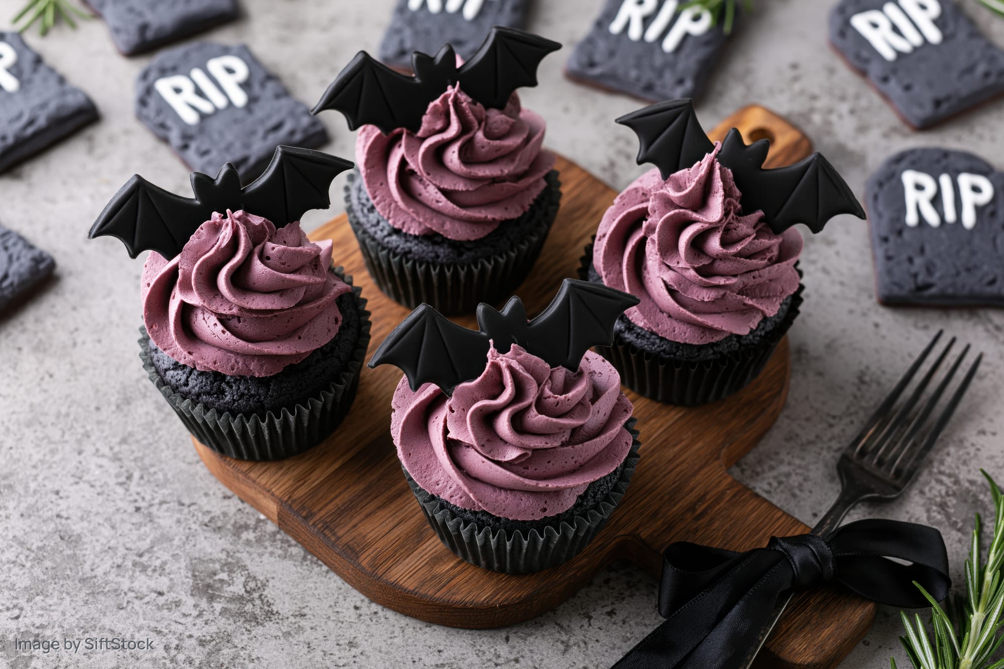 Halloween-themed chocolate cupcakes with purple frosting and bat wings on a wooden board surrounded by tombstone cookies