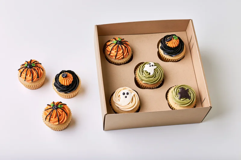 Halloween-themed cupcakes with pumpkin ghost and bat decorations arranged in a cardboard box and beside it on a white surface