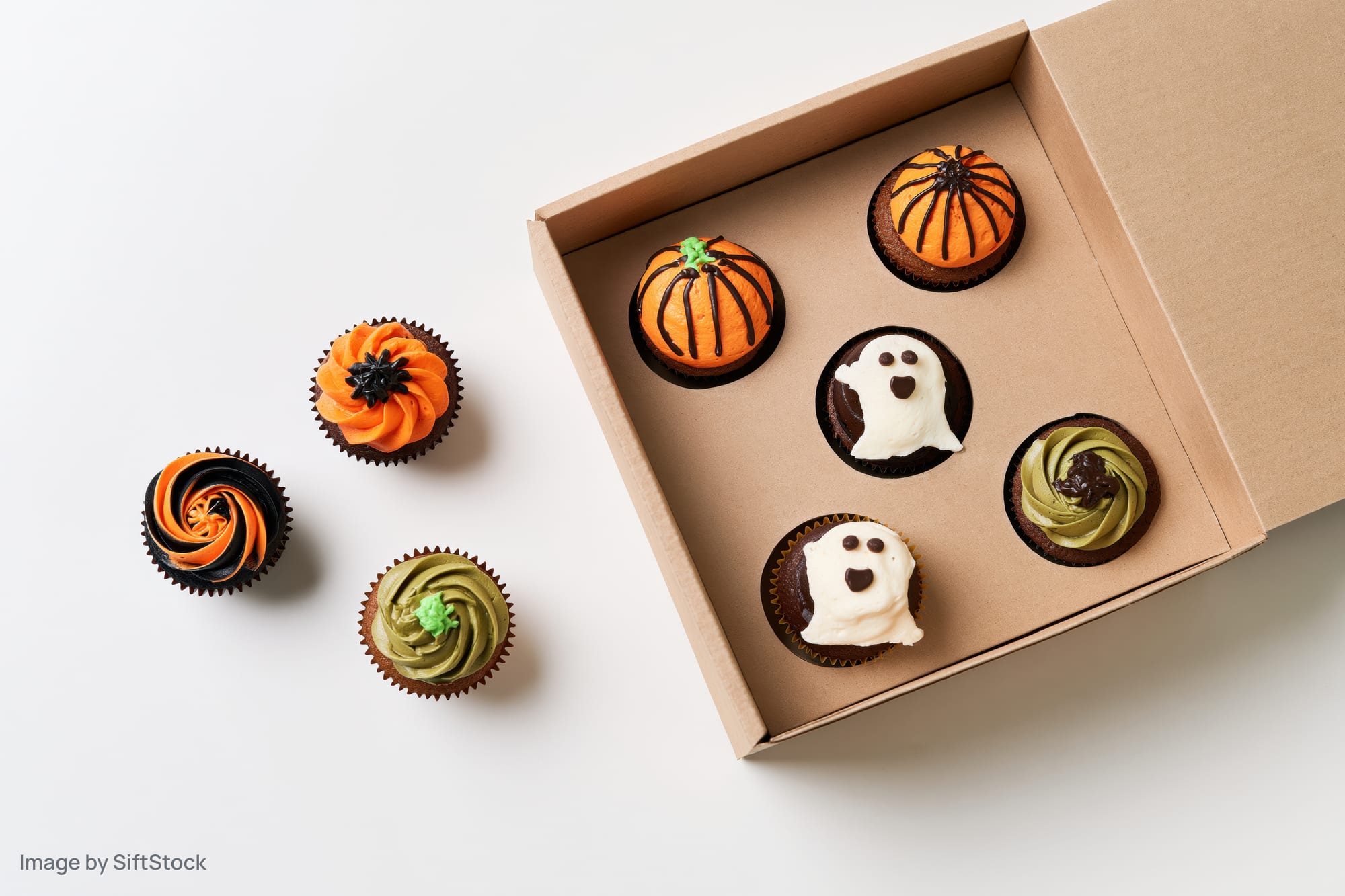 Halloween-themed cupcakes with pumpkin and ghost designs arranged in an open box and beside it on a white surface
