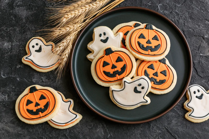 Halloween-themed sugar cookies shaped like jack-o-lanterns and ghosts on a dark plate with wheat stalks nearby