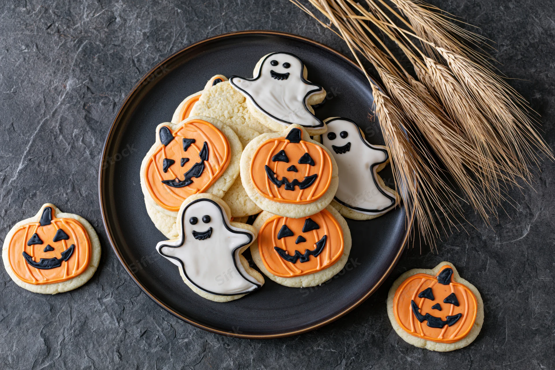 Halloween-themed sugar cookies shaped like jack-o-lanterns and ghosts on a black plate with straw broom beside on dark textured surface