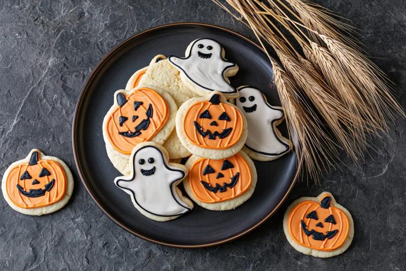 Halloween-themed sugar cookies shaped like jack-o-lanterns and ghosts on a black plate with straw broom beside on dark textured surface