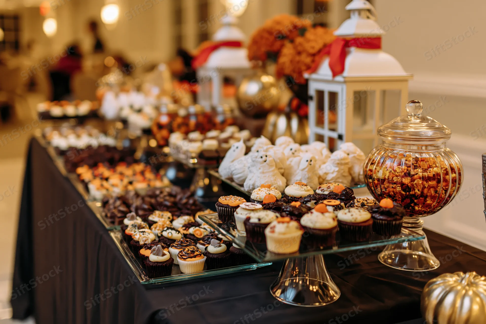Halloween-themed dessert table with decorated cupcakes and ghost meringues on a black cloth, surrounded by lanterns and pumpkin decor
