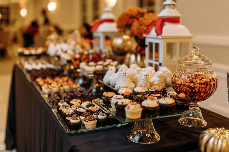 Halloween-themed dessert table with decorated cupcakes and ghost meringues on a black cloth, surrounded by lanterns and pumpkin decor