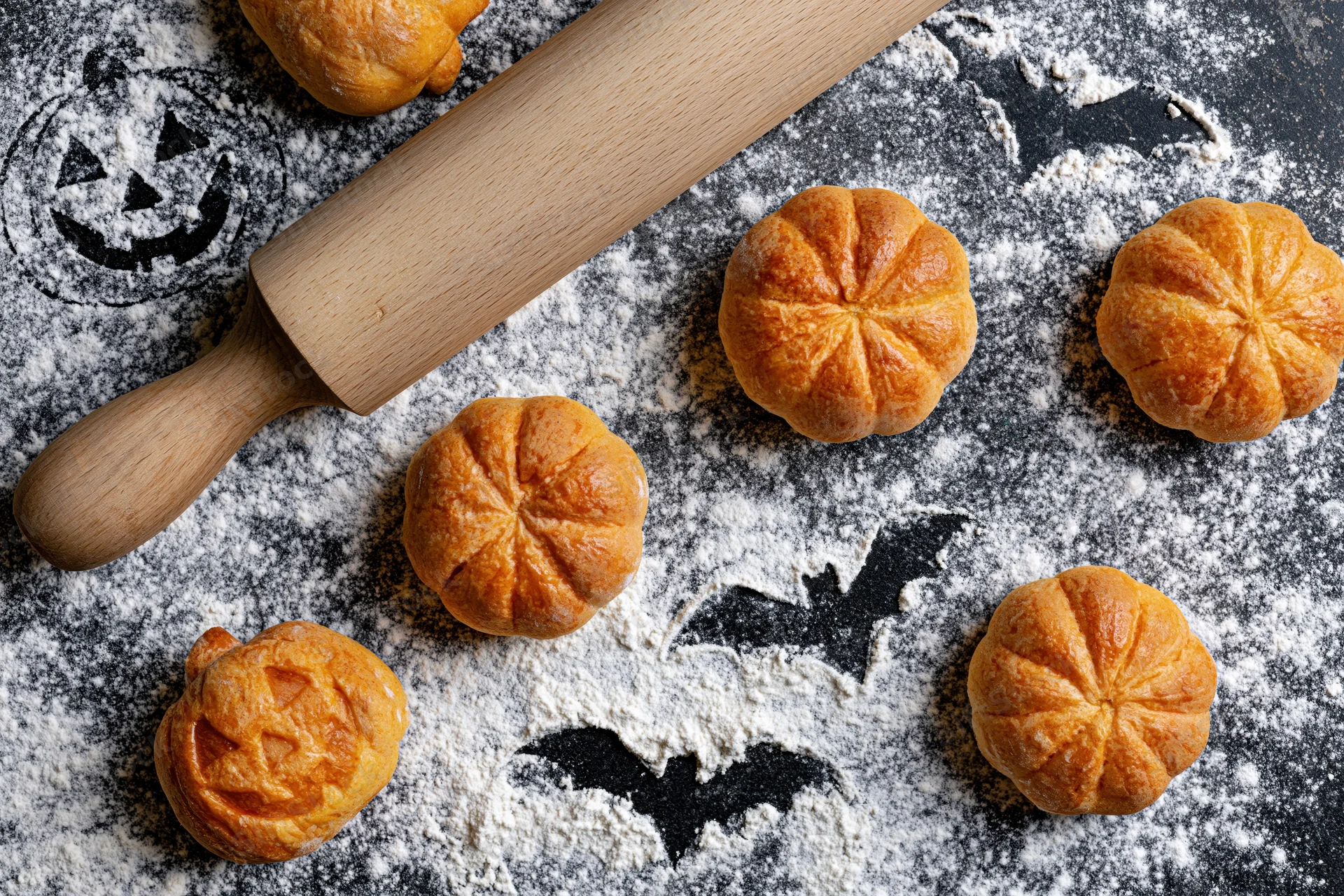 Pumpkin-shaped bread rolls and a wooden rolling pin arranged on a floured dark surface with halloween decorations