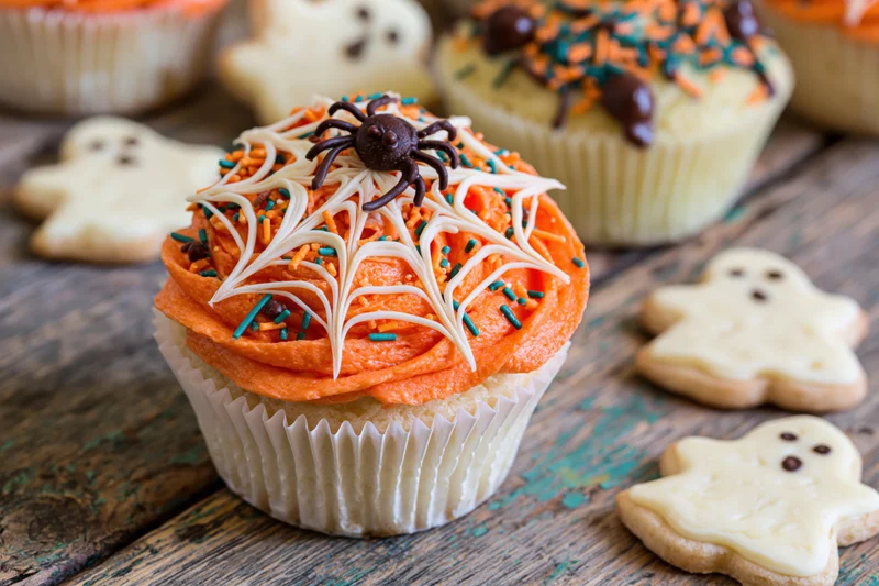 Halloween cupcake with orange frosting and spider web decoration on wooden table surrounded by ghost cookies