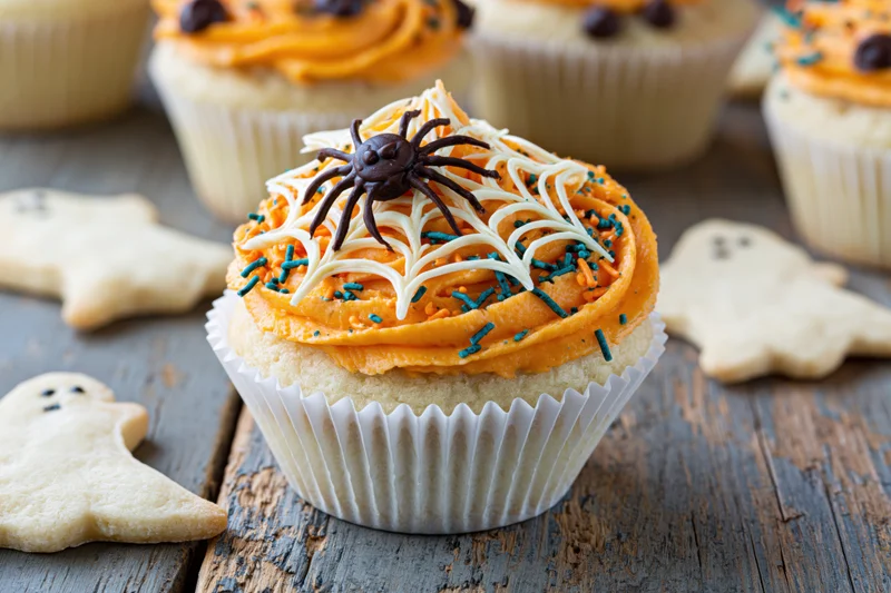 Halloween cupcake with orange frosting and chocolate spider on wooden table surrounded by ghost cookies