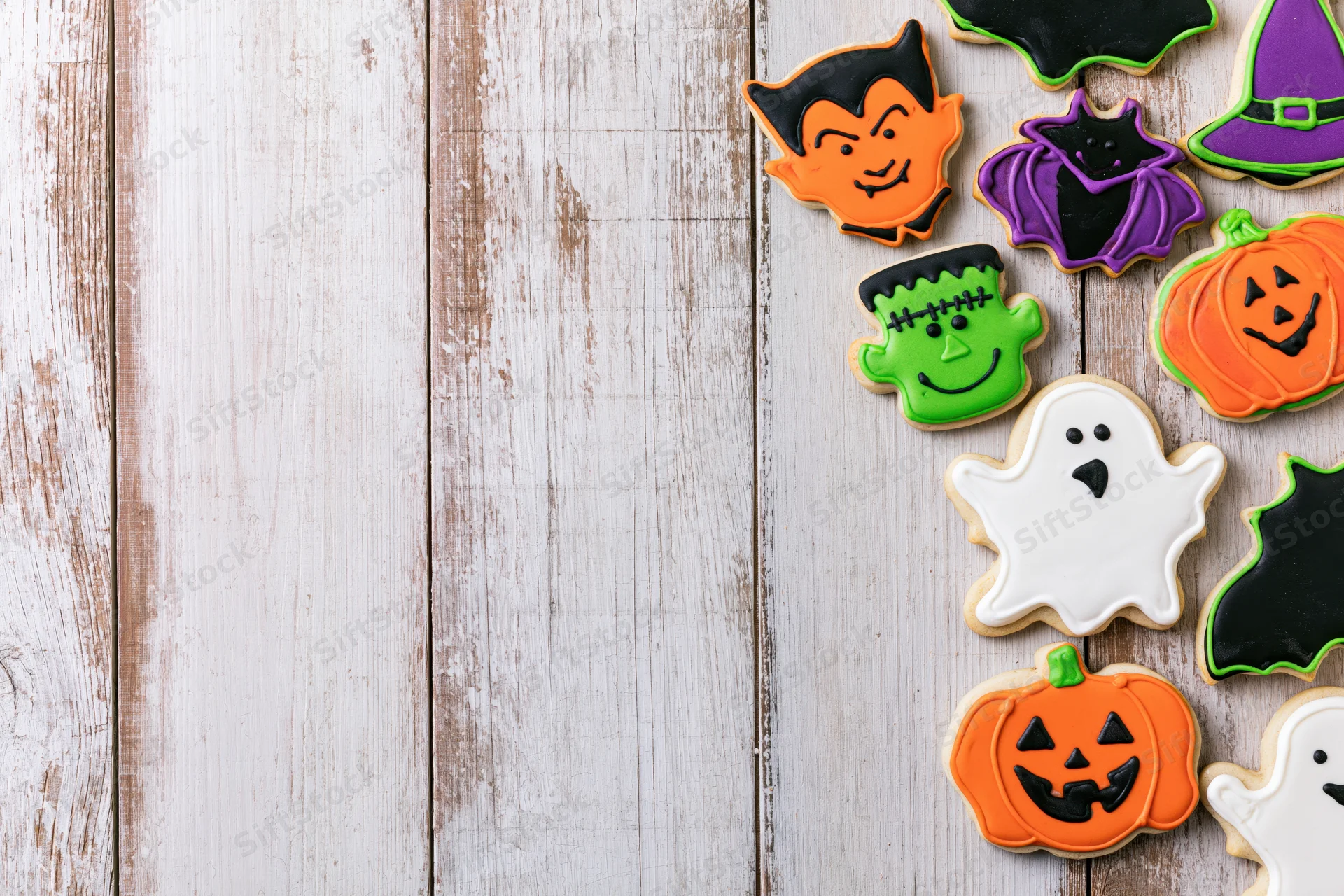 Halloween-themed sugar cookies shaped as ghosts, pumpkins, vampires, and witches arranged along the right side of a rustic white wooden table with copyspace on the left