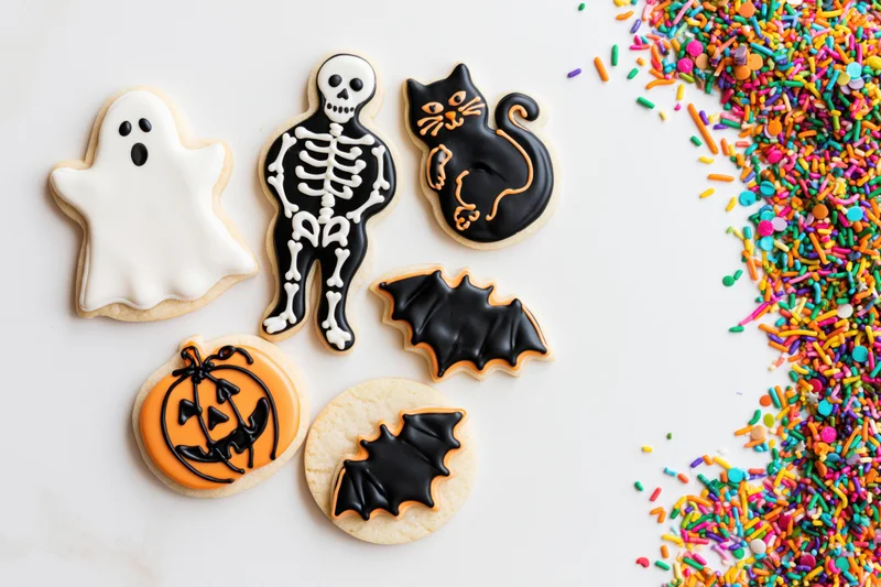 Halloween-themed sugar cookies shaped as ghost, skeleton, cat, bat and pumpkin with colorful sprinkles on white surface