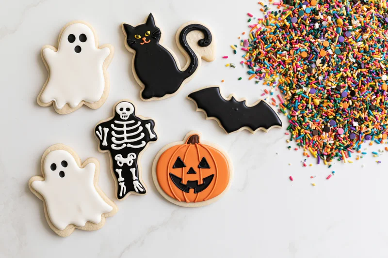 Halloween sugar cookies shaped as ghosts, cats, skeletons, pumpkins and bats arranged on a white marble surface with colorful sprinkles nearby