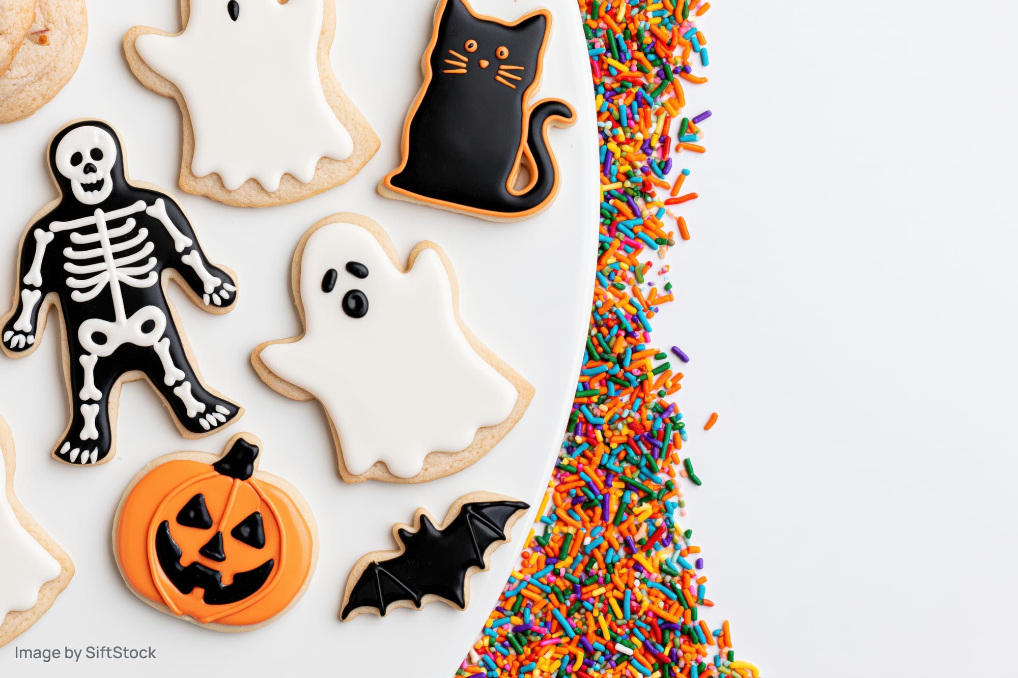 Halloween sugar cookies shaped as ghosts, skeletons, cats, pumpkins and bats on a white plate with colorful sprinkles beside it