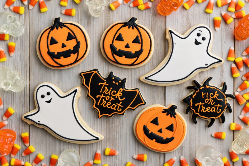 Halloween decorated sugar cookies shaped as pumpkins ghosts bats and spiders arranged on wooden surface with candy corn and plastic skulls