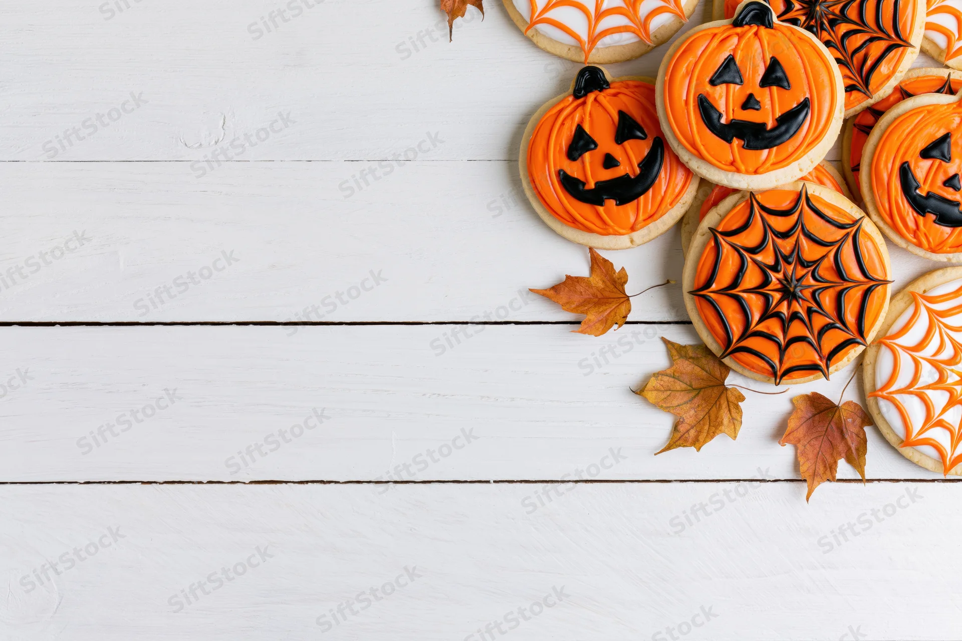 Halloween sugar cookies decorated with jack-o-lantern and spiderweb designs on white wooden surface with autumn leaves and copyspace