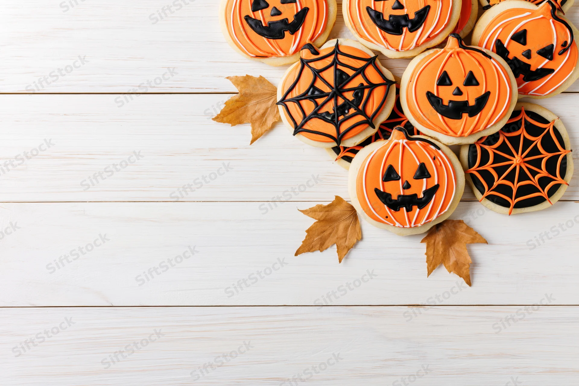 Halloween sugar cookies decorated with jack-o-lantern and spiderweb designs on white wooden surface with autumn leaves