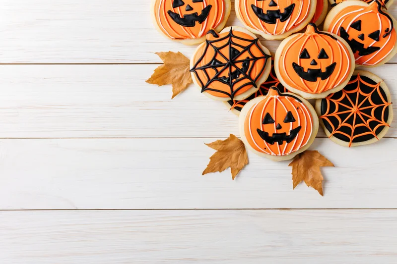 Halloween sugar cookies decorated with jack-o-lantern and spiderweb designs on white wooden surface with autumn leaves