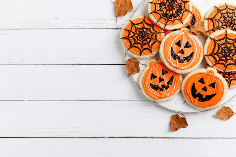 Halloween sugar cookies with jack-o-lantern and spiderweb designs on a white wooden table with autumn leaves and copyspace