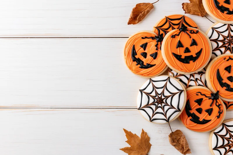Halloween sugar cookies decorated with jack-o-lantern faces and spiderwebs on white wooden surface with dried autumn leaves