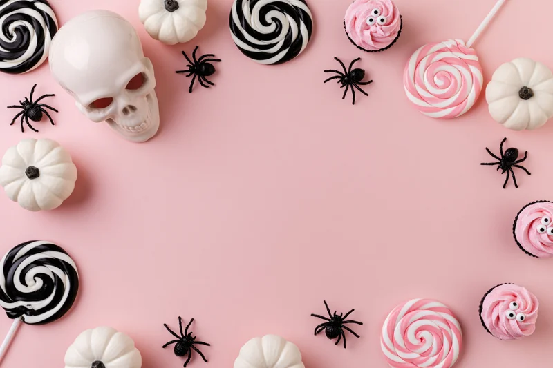 Halloween flat lay with candy, skull, pumpkins and spiders arranged around pink background with copyspace