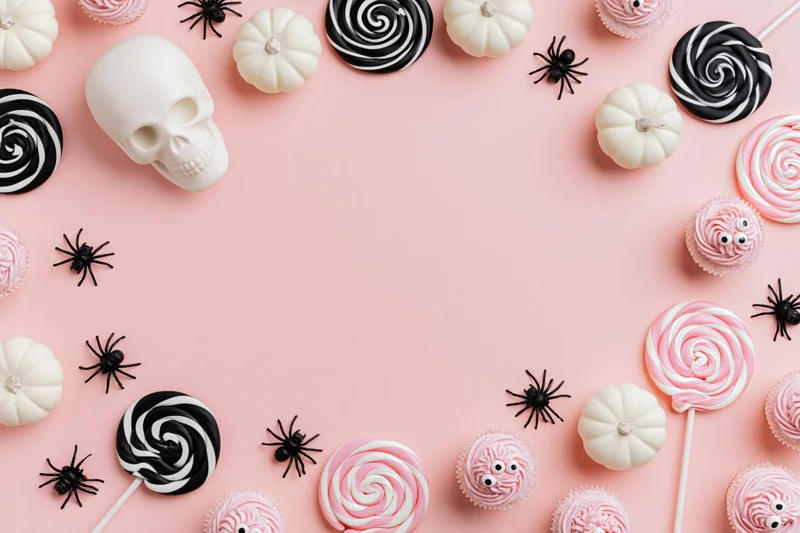 Halloween flat lay with candy, skull, pumpkins and spiders arranged around pink background with copyspace