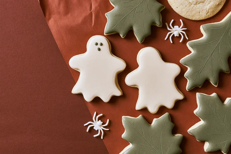 Halloween sugar cookies shaped as ghosts and leaves with plastic spiders on rust-brown paper