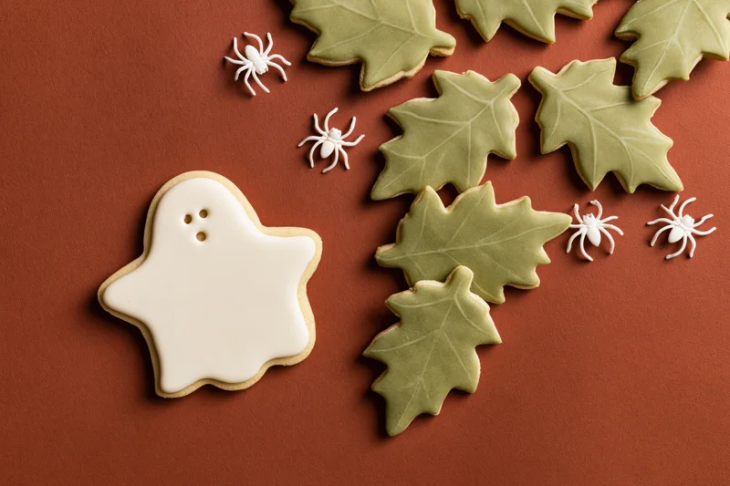 Halloween sugar cookies shaped as ghosts and leaves with white spiders arranged on rust-brown paper background