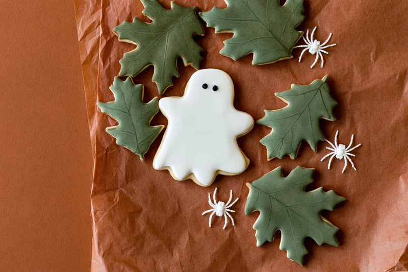 Halloween sugar cookies shaped as ghosts and leaves with white spiders arranged on crumpled brown paper