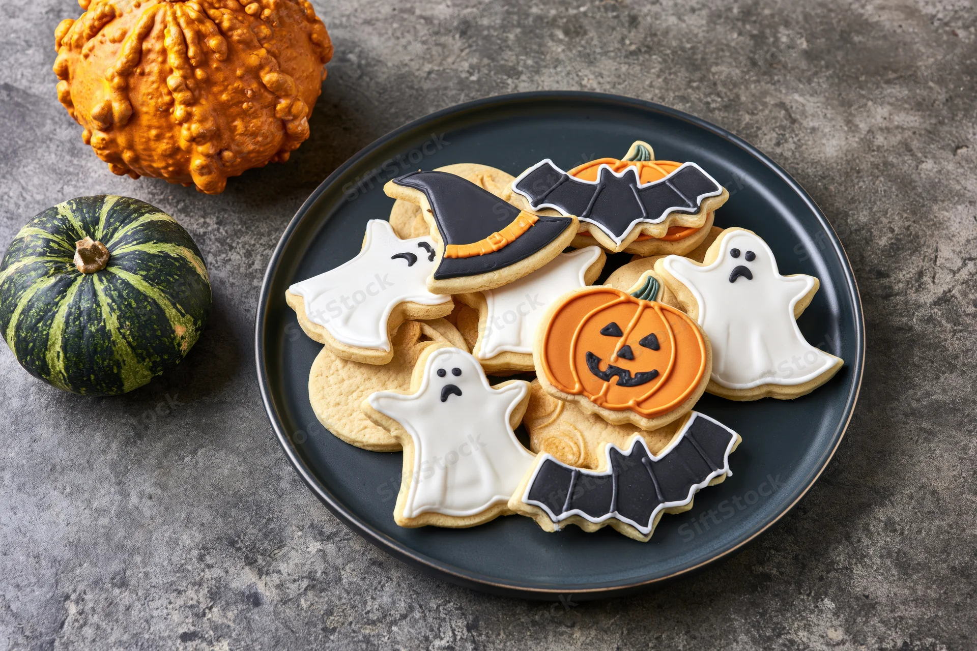 Halloween sugar cookies decorated with royal icing in ghost pumpkin bat and witch hat shapes on a black plate with pumpkins beside it