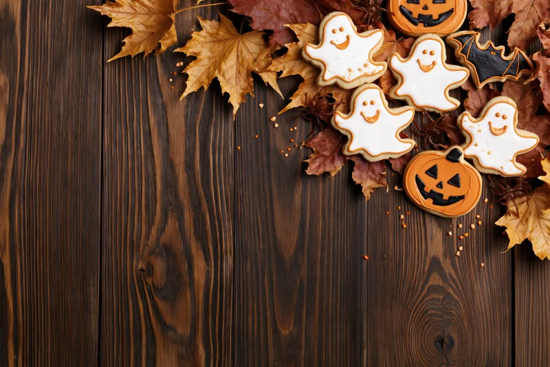 Halloween sugar cookies shaped like ghosts and jack-o-lanterns arranged with autumn leaves on a dark wooden table