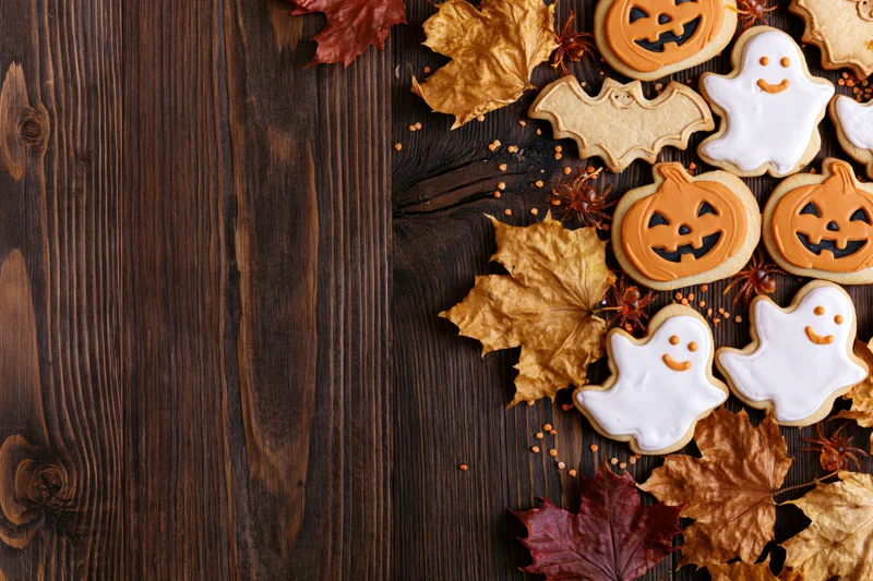 Halloween sugar cookies shaped like ghosts and jack-o-lanterns arranged with autumn leaves on a dark wooden table