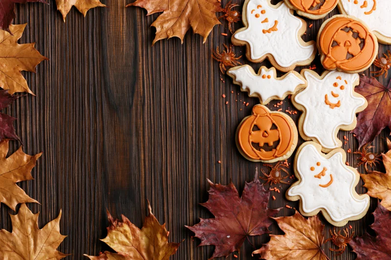 Halloween sugar cookies shaped like ghosts and jack-o-lanterns arranged with autumn leaves on a dark wooden surface