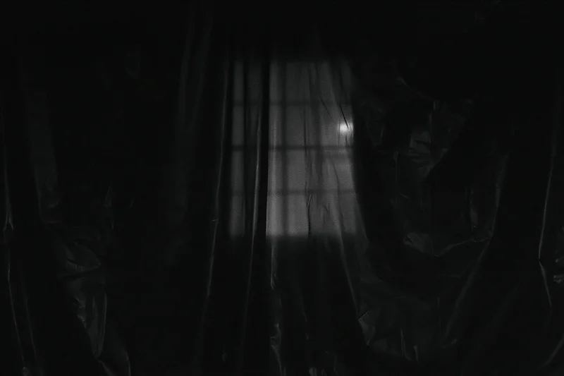 Dark plastic sheeting hanging in vertical folds inside a dimly lit space with light filtering through a grid-patterned window